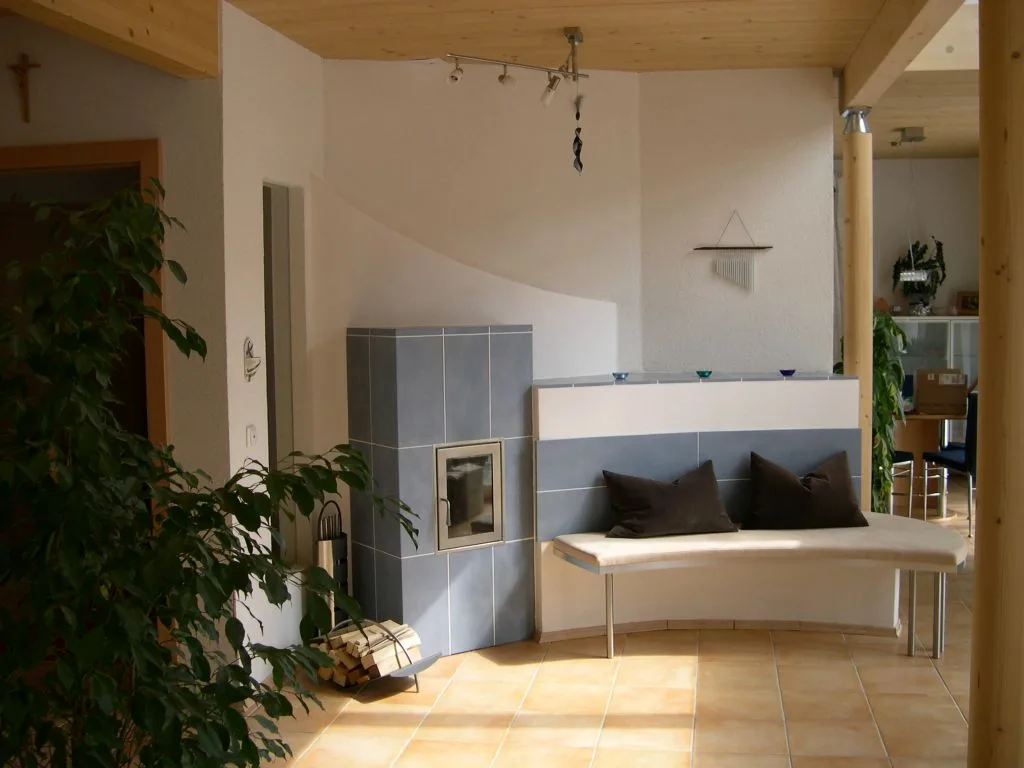Graublau gefliester Kachelofen mit geschwungener Sitzbank und Kissen unter einer Holzdecke